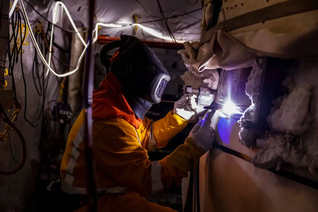 A worker wearing protective gear and a welding helmet uses a welding torch on a metal surface in an industrial setting, surrounded by hanging cables and insulated materials. Blue sparks illuminate the scene.