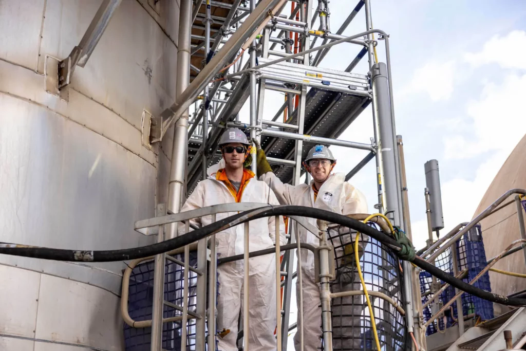 Two workers in white protective suits and helmets stand on an industrial scaffold platform, surrounded by pipes and metal structures, with one worker holding onto a railing. Blue sky is visible in the background.