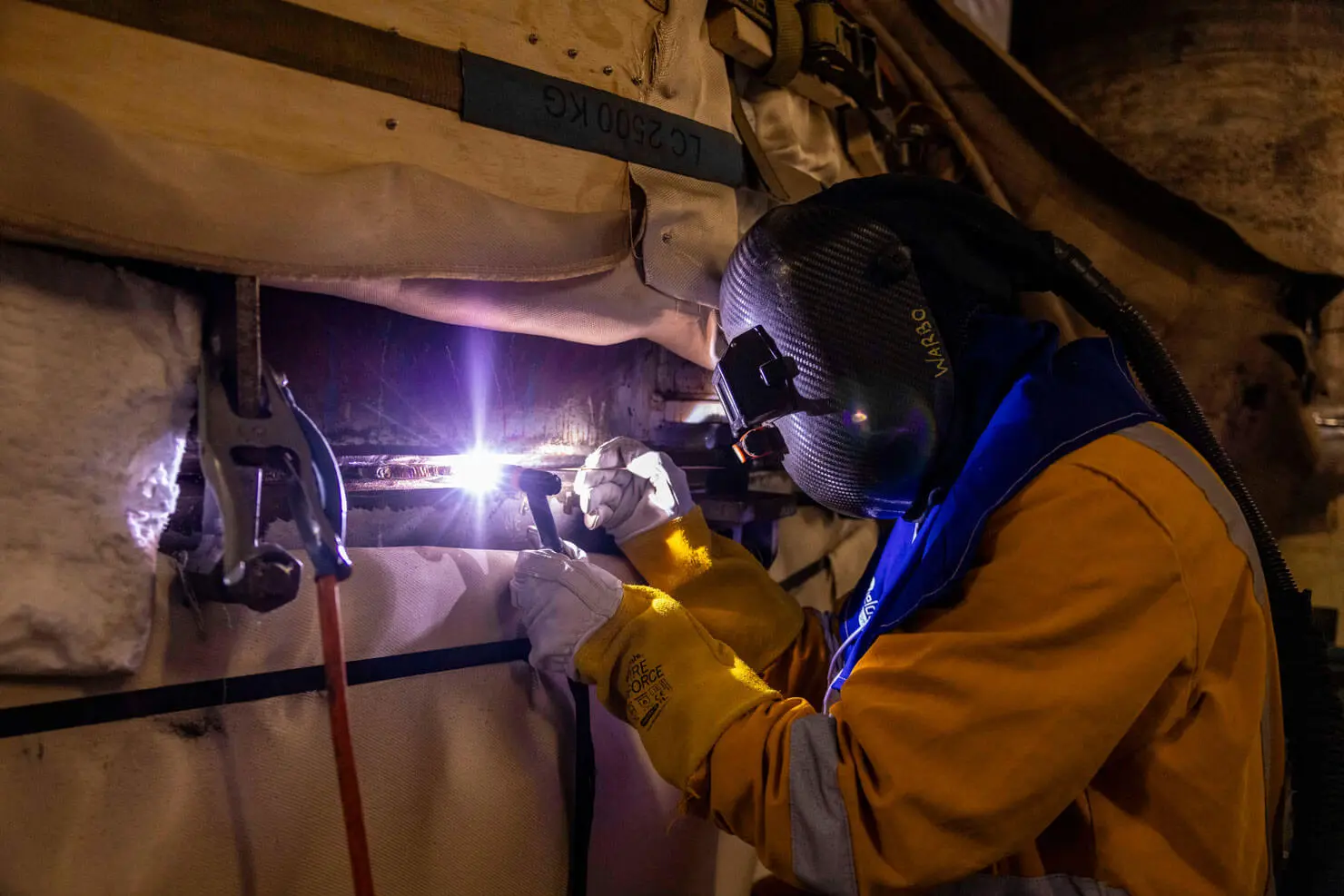 A worker in protective gear and mask welds metal, creating bright sparks, inside an industrial setting with insulated pipes and clamps visible.