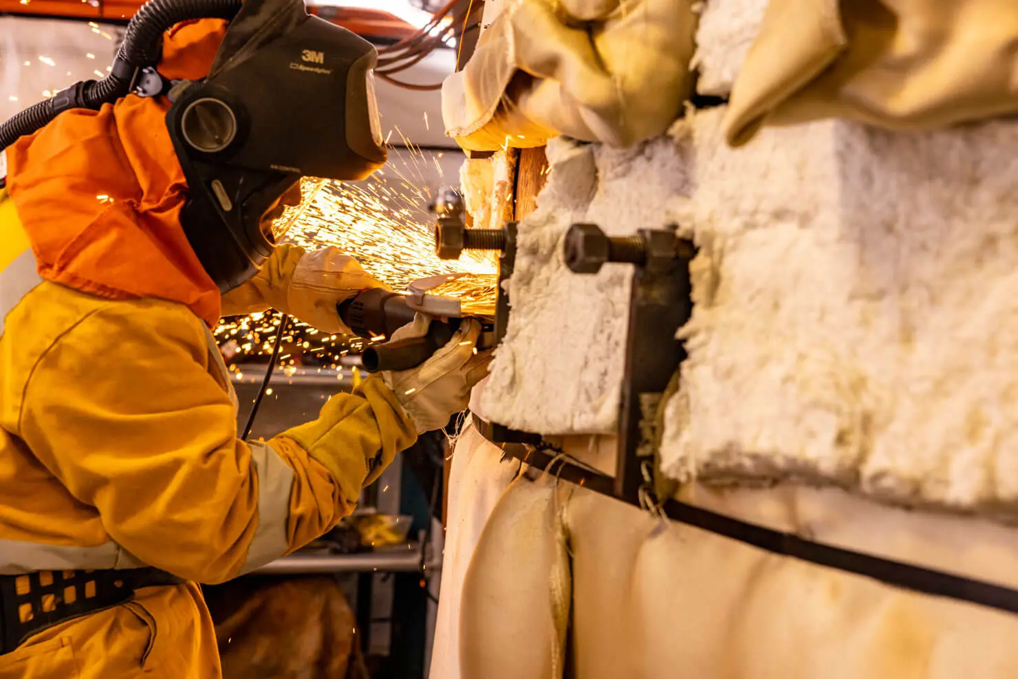 A worker in protective gear uses a power tool to cut metal, creating a shower of sparks. Insulated pipes or equipment are visible beside the worker in an industrial setting.