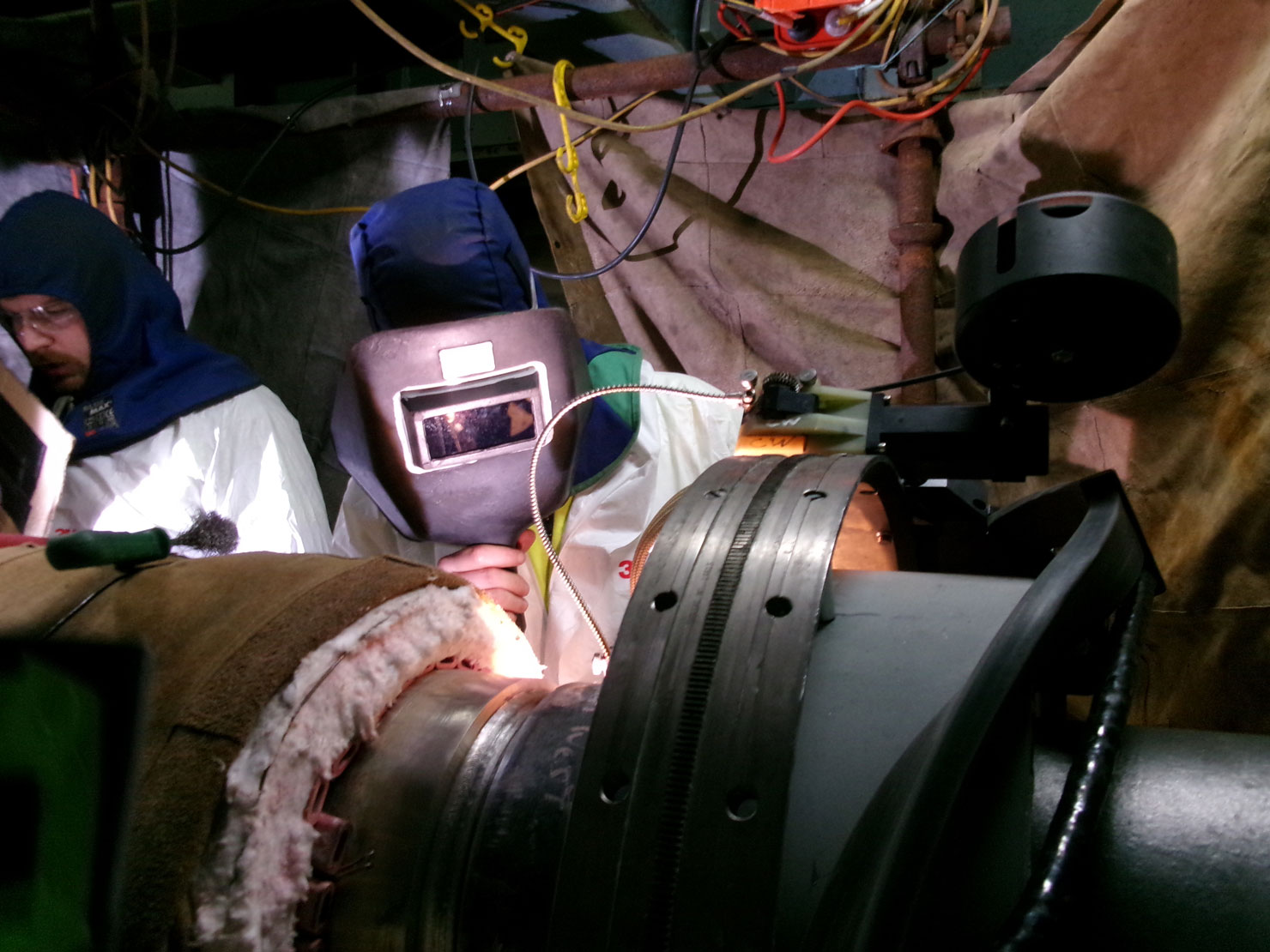 Two workers in protective clothing and welding masks perform welding on a large metal pipe in an industrial setting with wires, tools, and equipment visible around them.