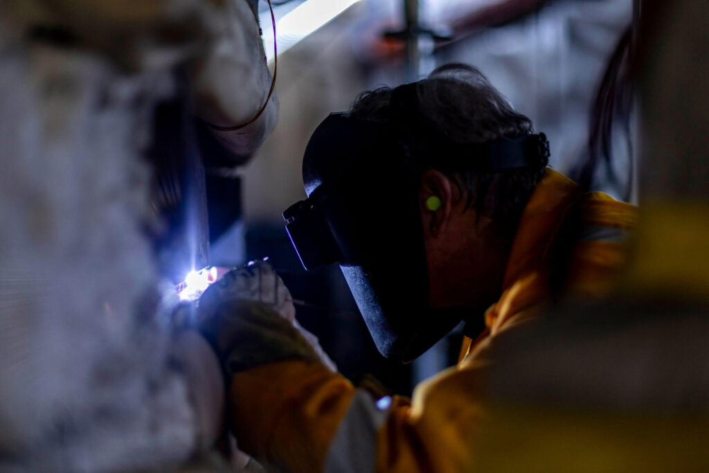 A person wearing protective gear and a welding mask is working with bright sparks in a dimly lit industrial setting.