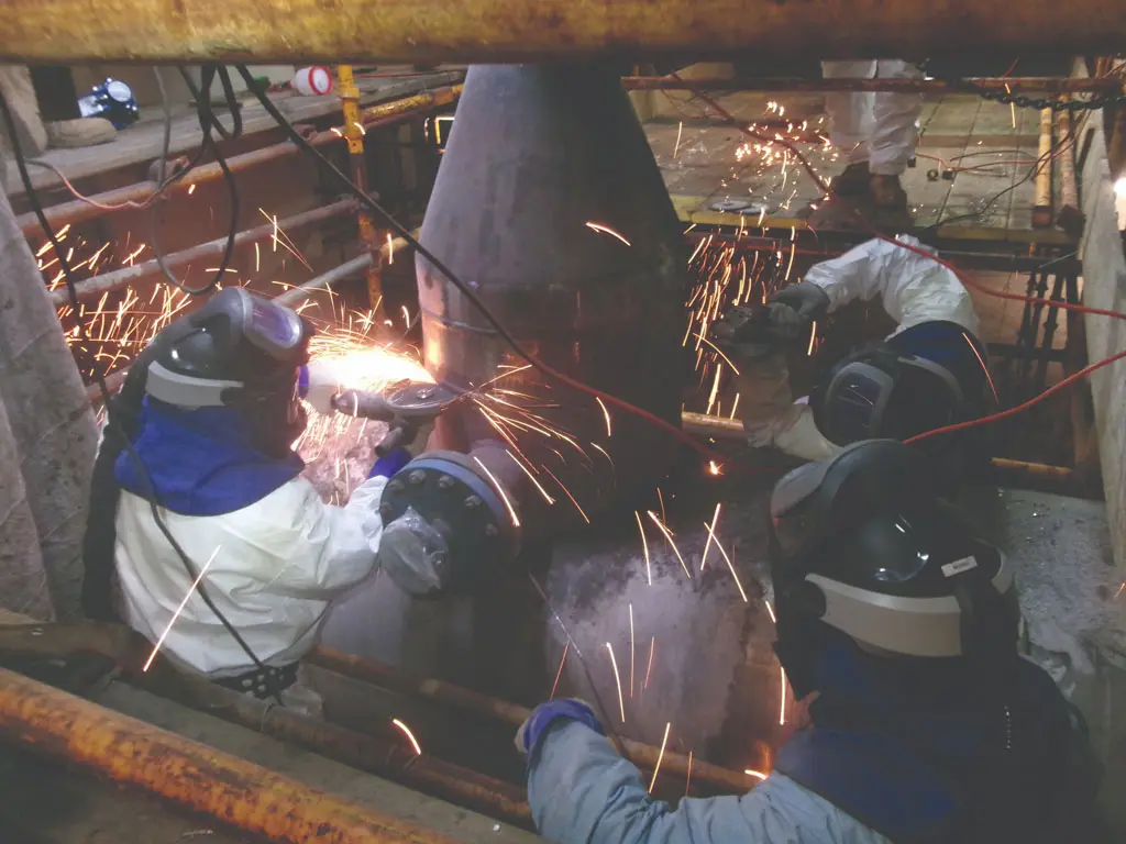 Three workers in protective gear use power tools to cut or grind a large metal structure, with sparks flying around them in an industrial setting surrounded by pipes and scaffolding.