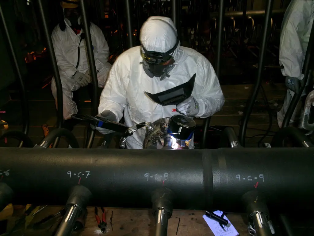 A worker in protective white coveralls and goggles welds a metal pipe in an industrial setting, surrounded by equipment and tools. Numbers are marked in white on the black pipes.