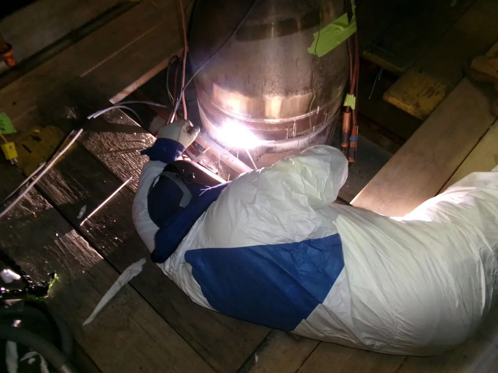 A person in a white protective suit with blue patches is welding or working on a large metal cylinder, surrounded by cables, on a wooden platform. Bright light is visible at the work site.