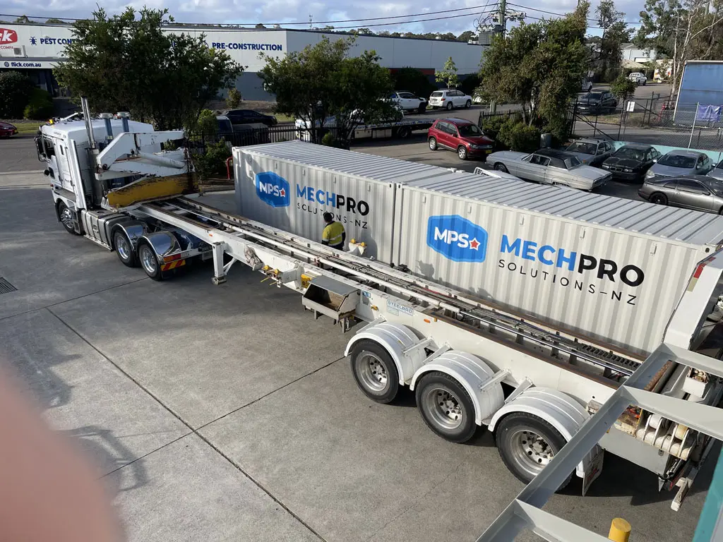 A white truck with a crane is transporting two large MECHPRO Solutions-NZ shipping containers on a long trailer in an industrial parking lot. Several cars and trees are visible in the background.