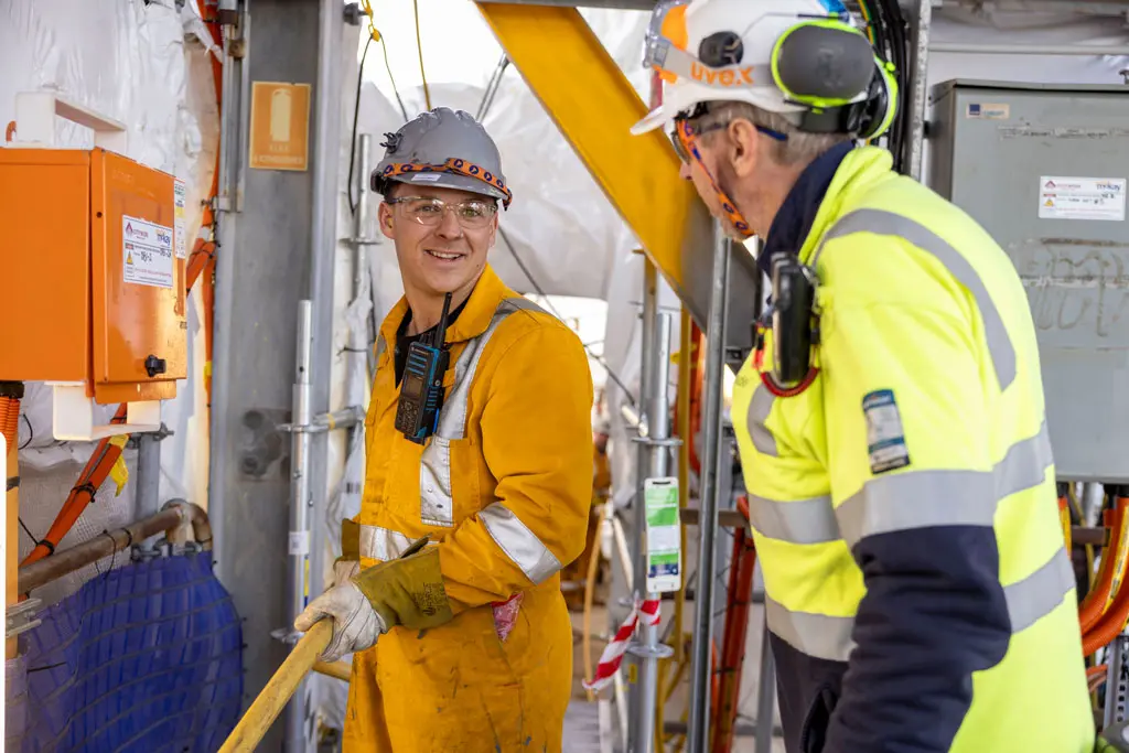 Two construction workers wearing safety gear and helmets talk inside a worksite. One in a yellow jumpsuit holds a tool, while the other in a high-visibility jacket faces him. Equipment and cables are visible around them.